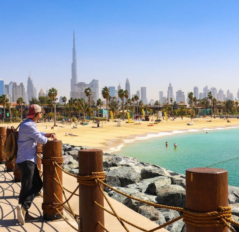 Mann lehnt an einem Holzgeländer mit Blick auf einen Strand und die Skyline von Dubai City.