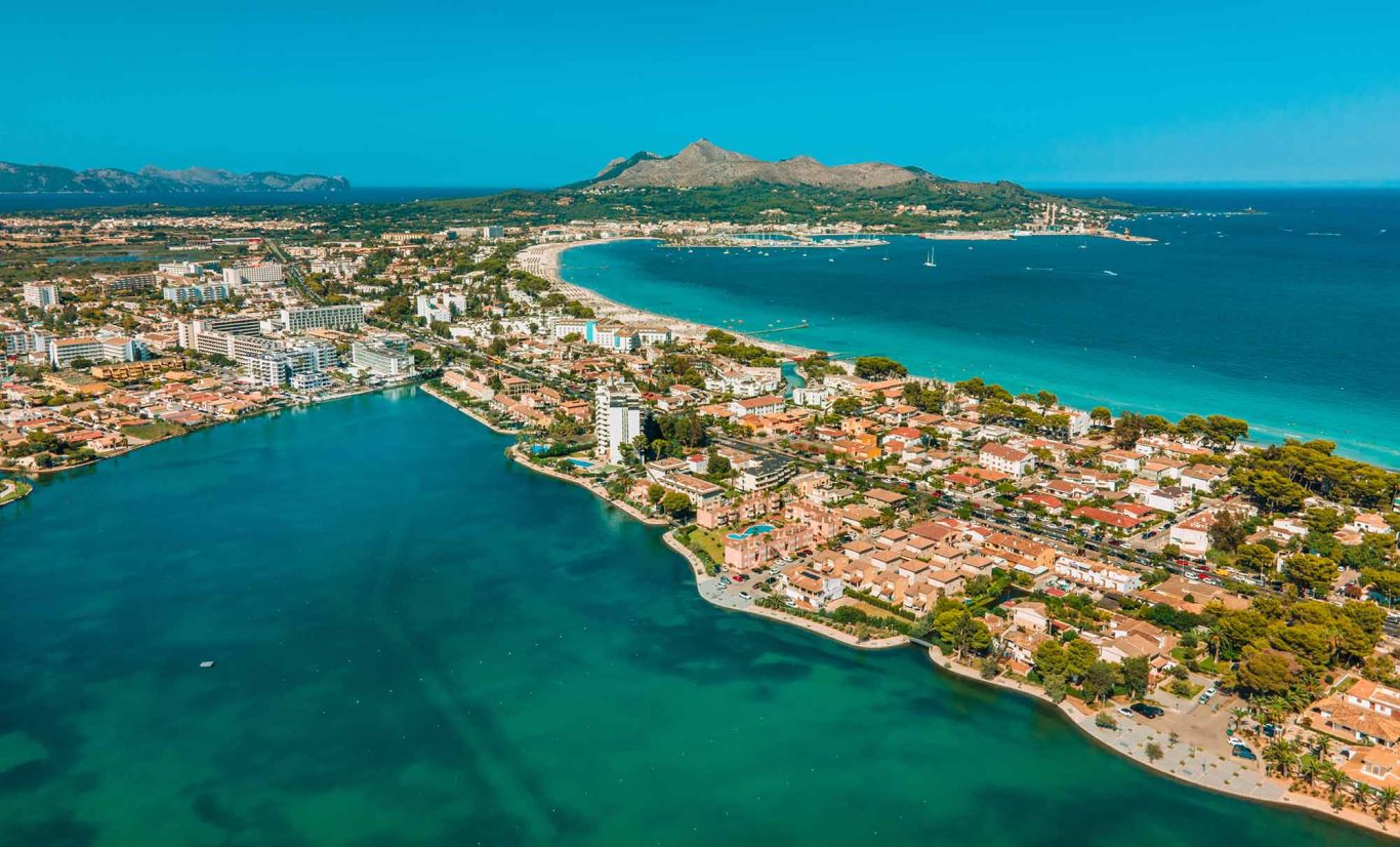 Alcúdia aus der Möwen-Perspektive mit Hotels am Strand Luftaufnahme einer Küstenstadt mit klaren Gewässern und Bergen im Hintergrund.