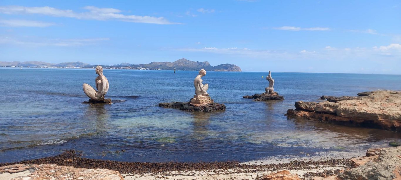 Berühmte Skulpturen am Strand in Can Picafort Drei Skulpturen stehen auf Steinen im ruhigen Wasser eines Strandes bei klarem Himmel.