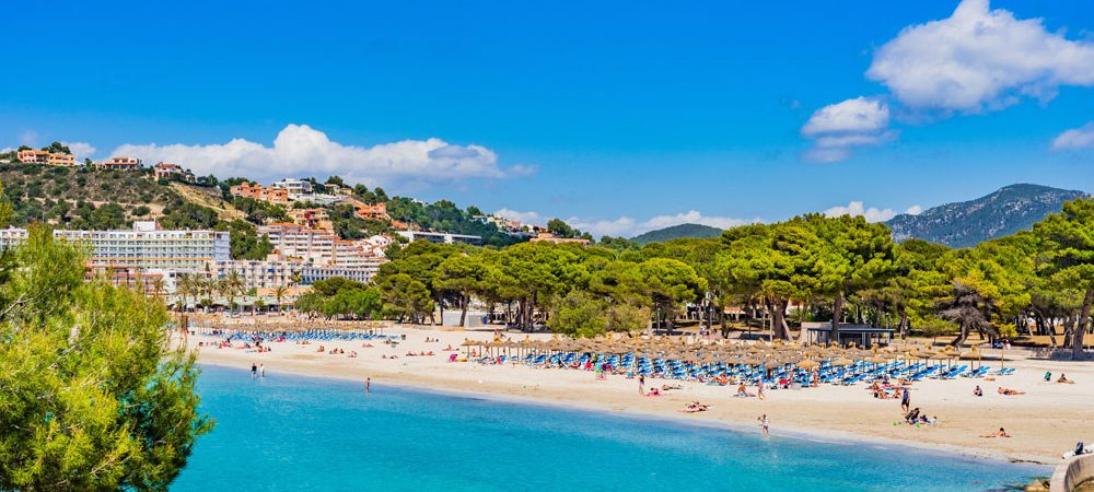 Klarer Himmel über einem Strand mit Palmen und Sonnenliegen, Menschen entspannen am Wasser.