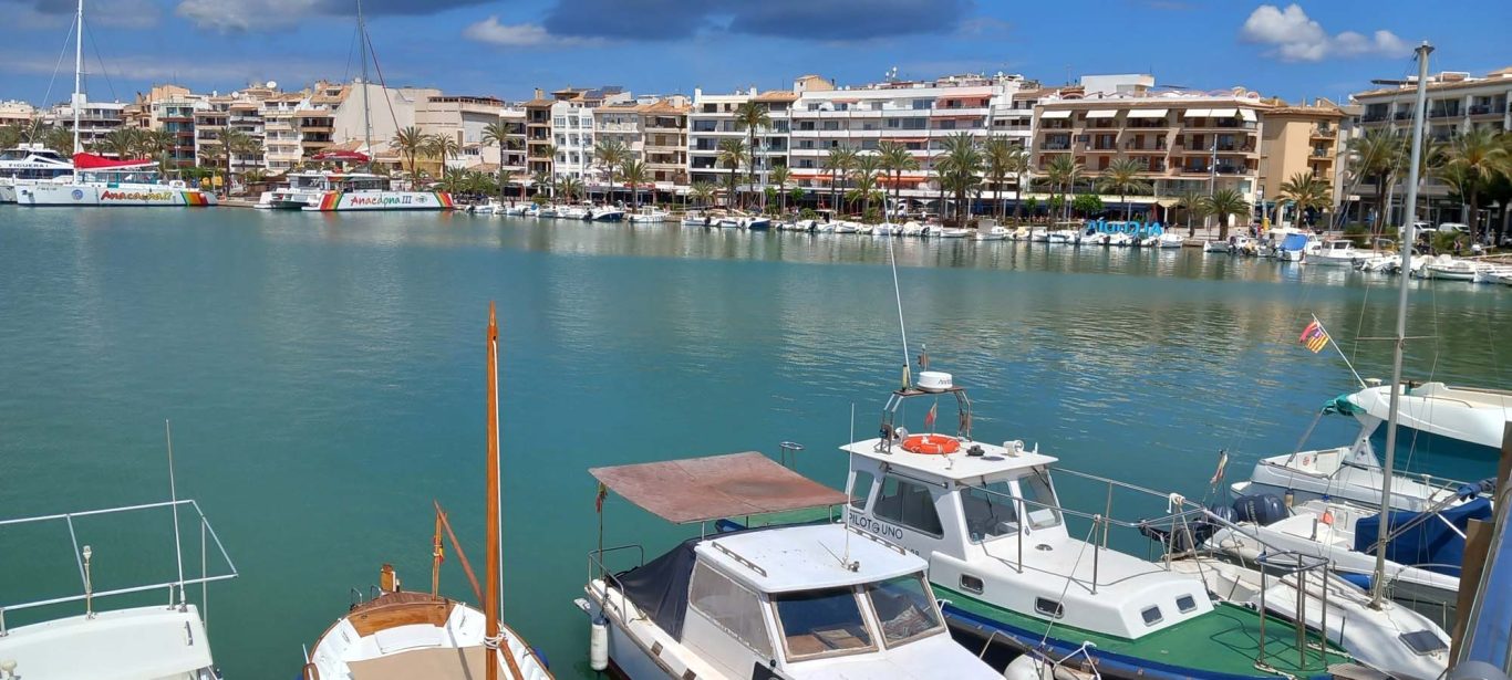 Hafenpromenade am Hafen in Alcúdia Blick auf einen Hafen mit Booten und einer Stadtansicht im Hintergrund.