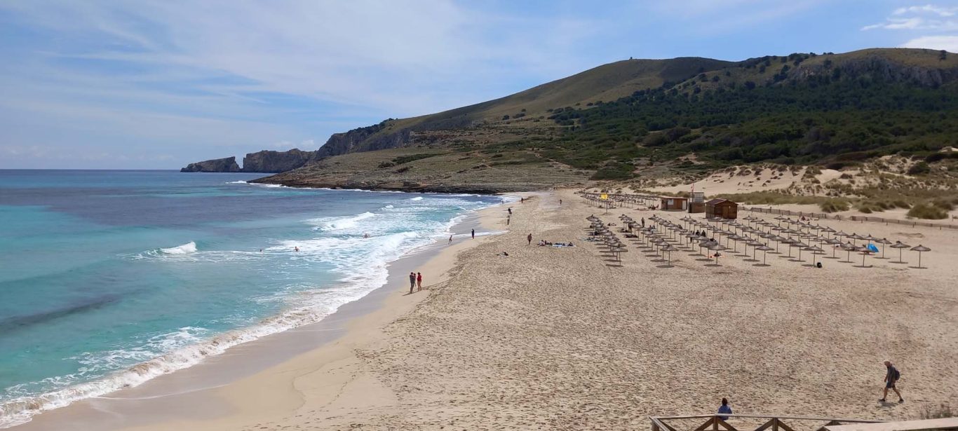 Strand in der Cala Mesquida 20 km östlich von Alcúdia Strand mit Wellen, sandigem Ufer und Bergen im Hintergrund. Sonniger Himmel.